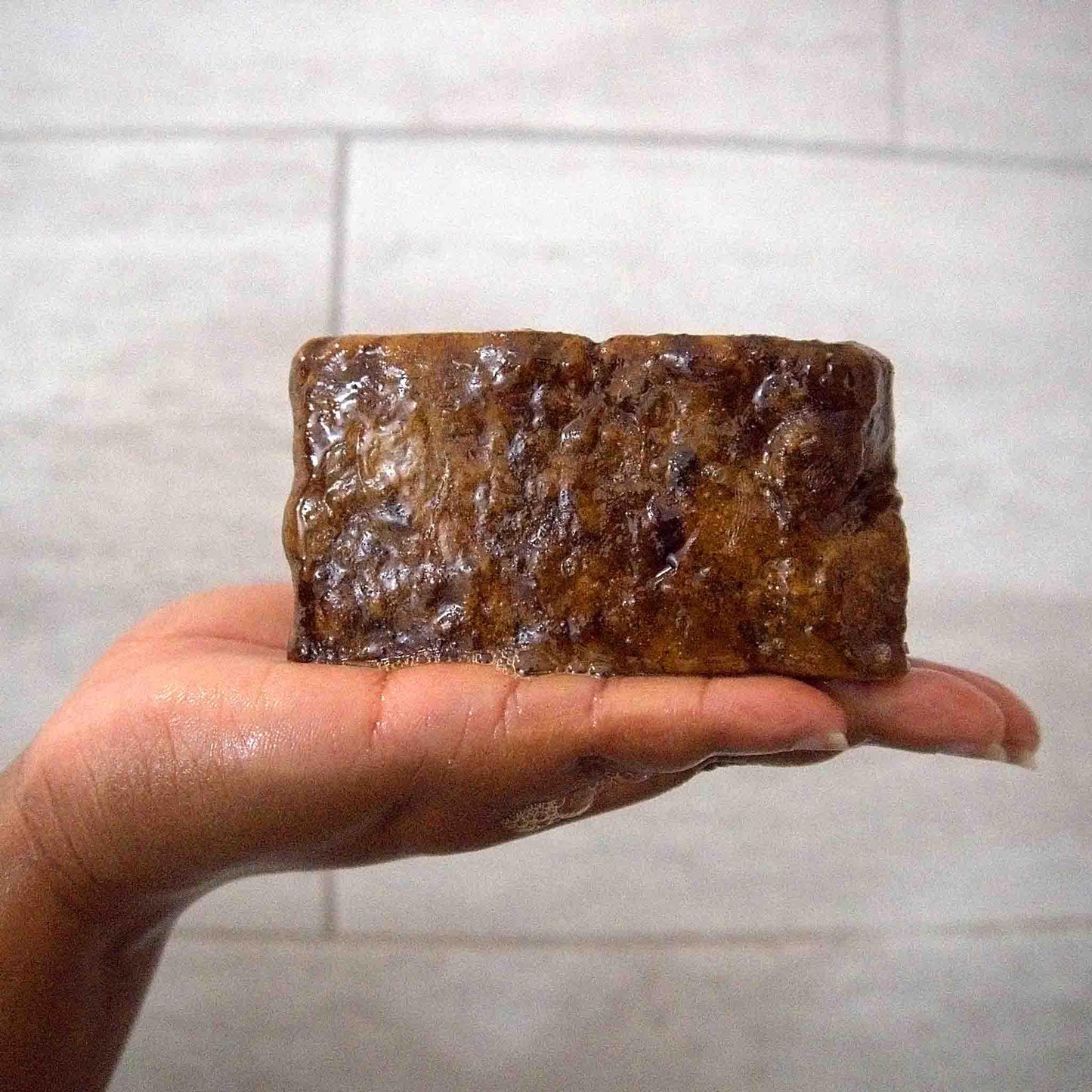 Hand holding wet, textured brown soap bar against tiled bathroom wall
