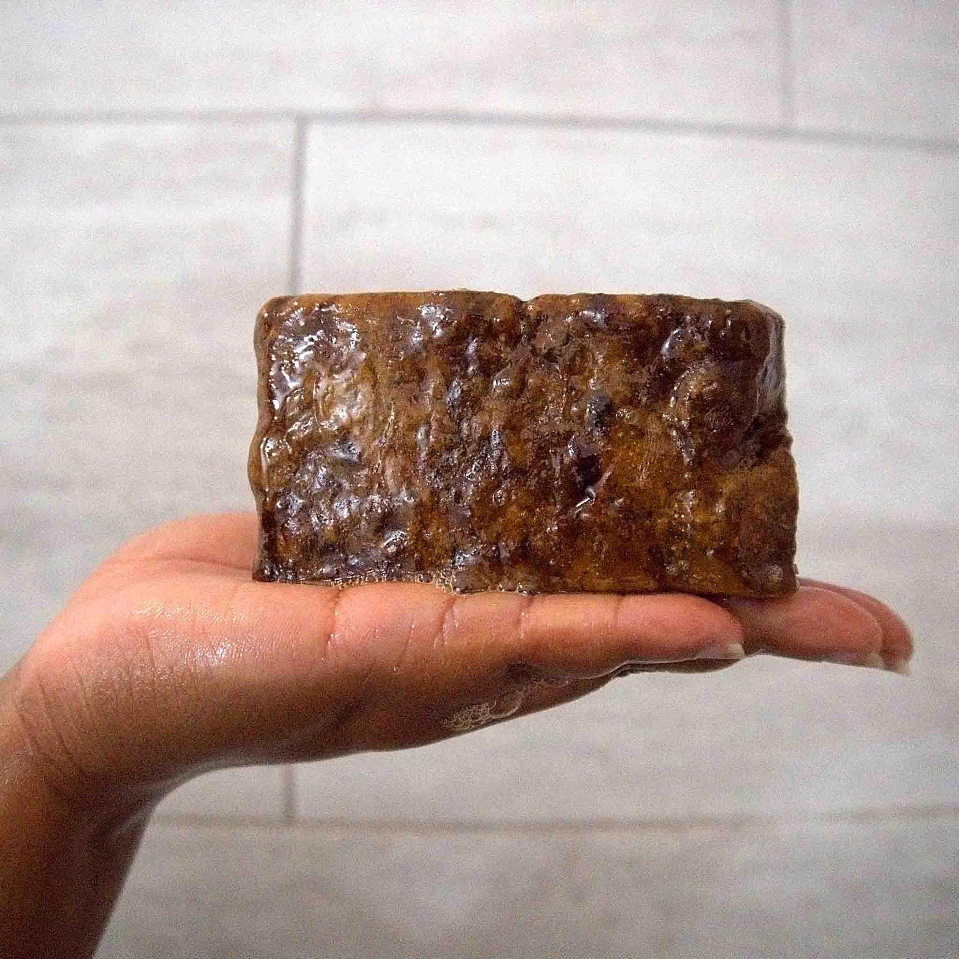Hand holding wet, textured brown soap bar against tiled bathroom wall