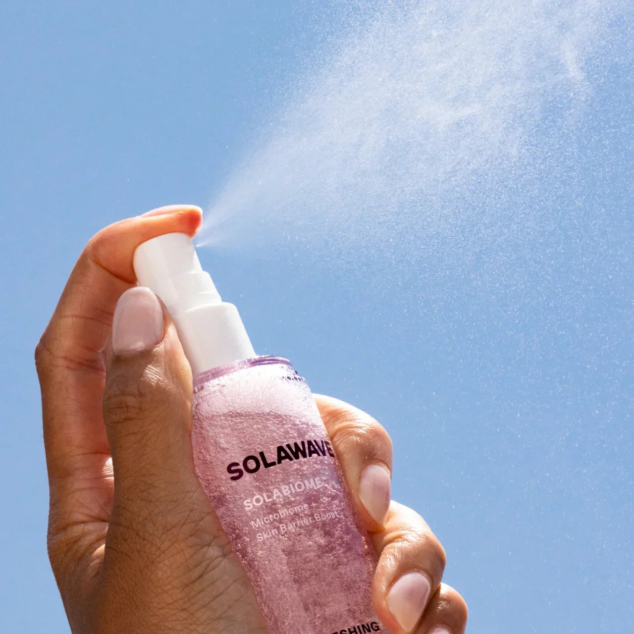 Hand holding Solawave spray bottle misting pink skincare solution against blue sky