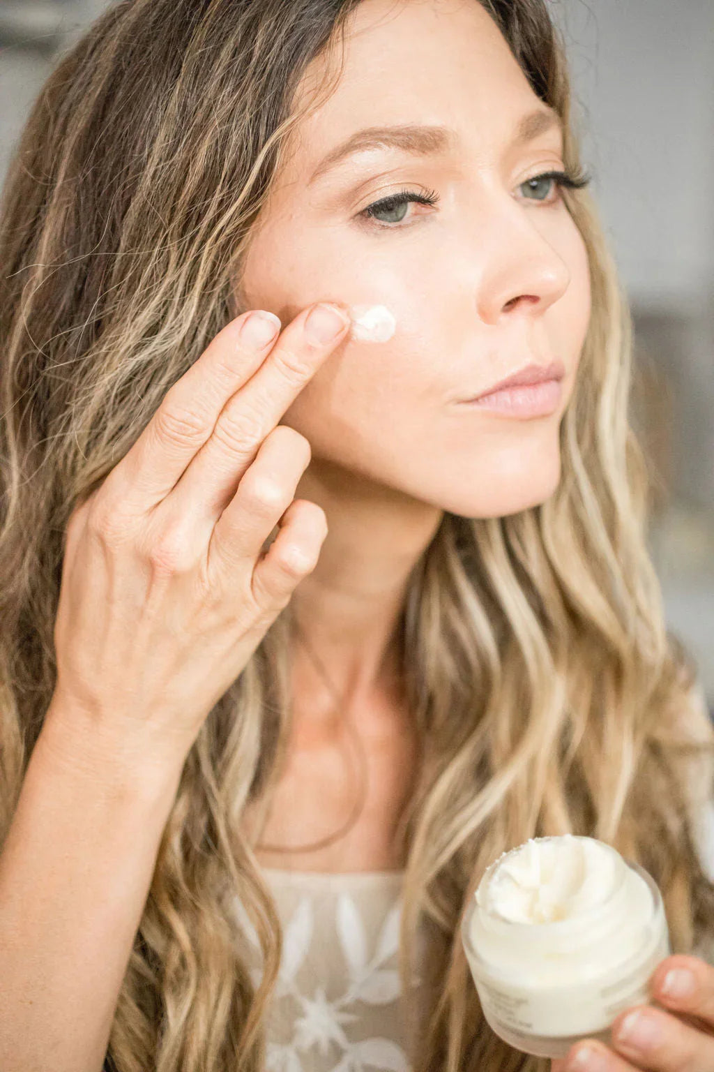 Woman applying hydrating face cream to cheek, holding jar of moisturizer for glowing skin routine
